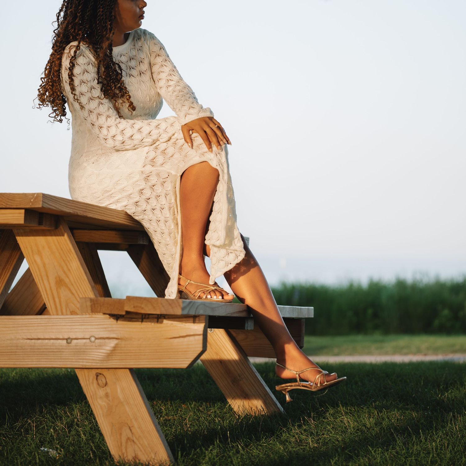 Woman in white lace dress sitting on wooden picnic table outdoors on green lawn at dusk.