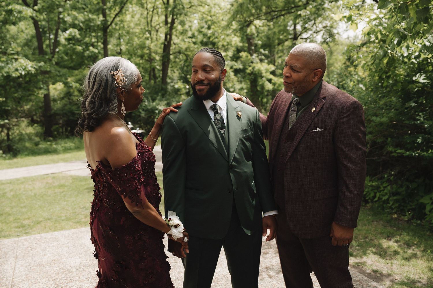 A groom in a green suit smiles with his parents outdoors at a wedding celebration.