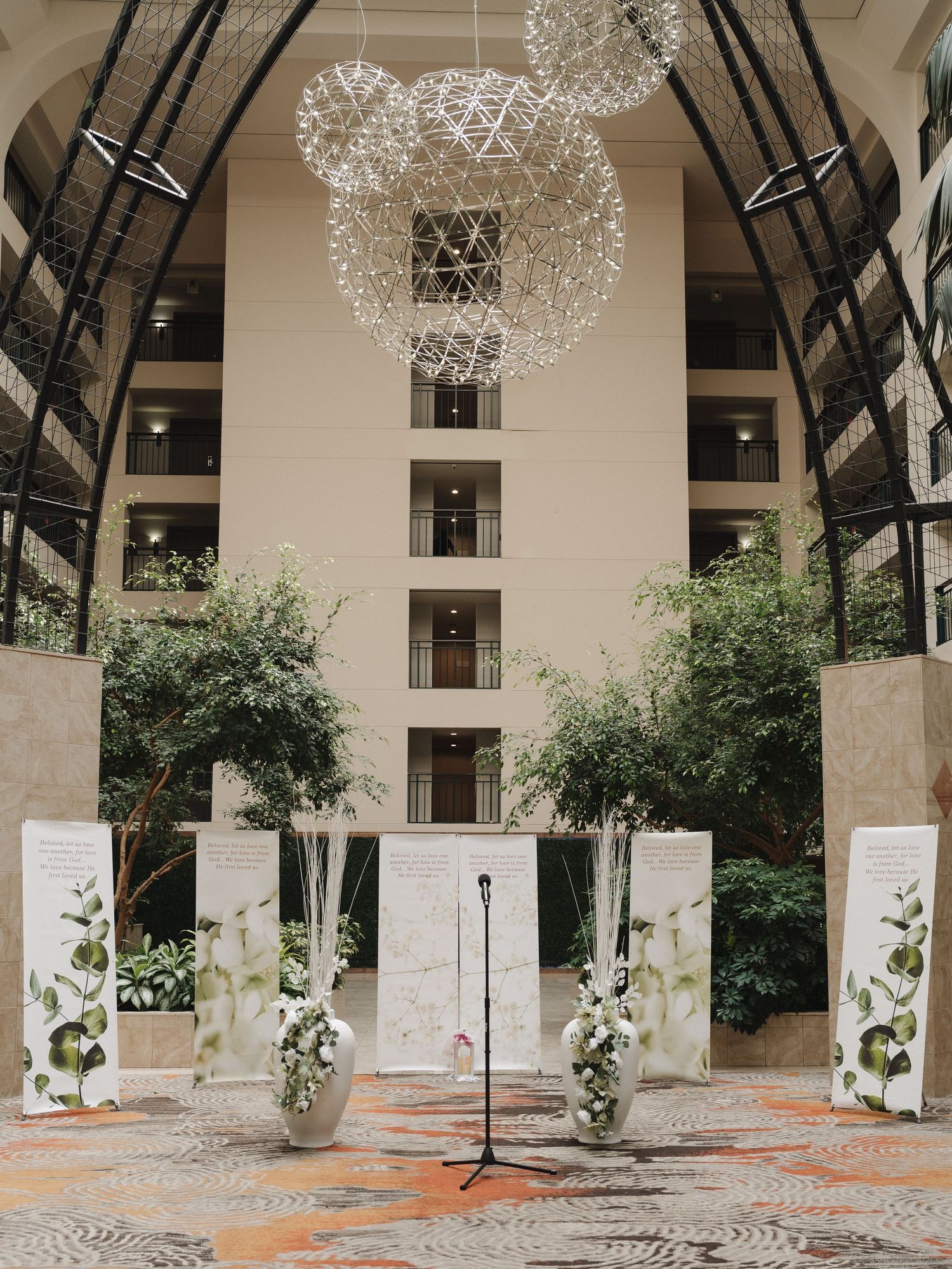 Elegant hotel atrium wedding ceremony setup with floral arrangements, white panels, and modern geometric chandeliers.