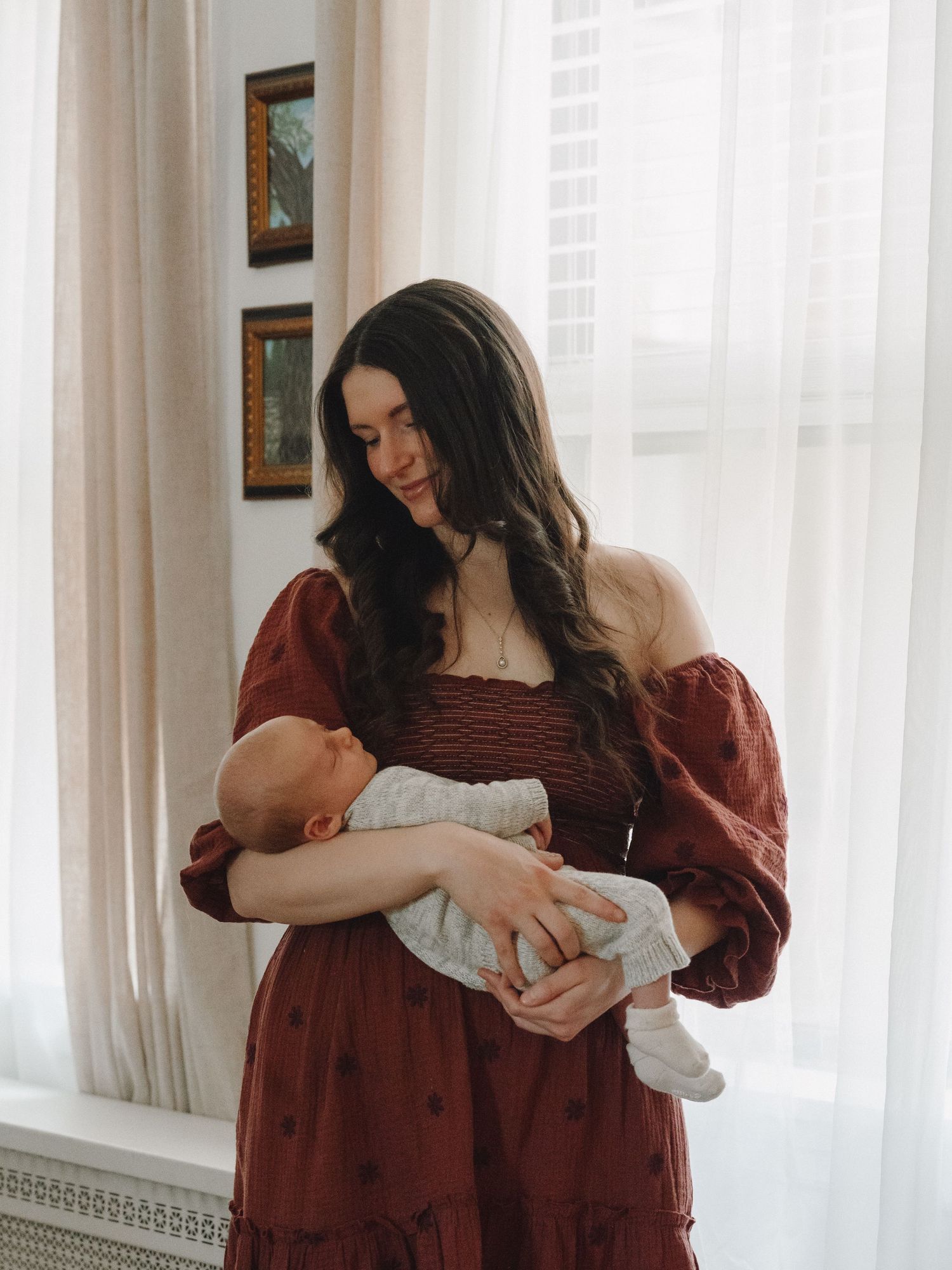 Young woman in red dress tenderly holds newborn baby near bright window indoors.
