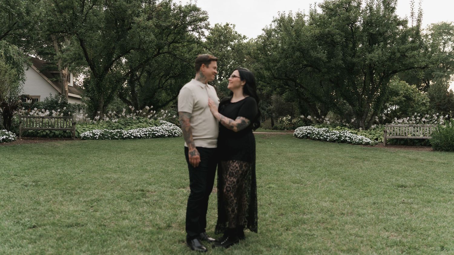 A couple stands together on a lush green lawn surrounded by trees and white flowers in a garden setting.