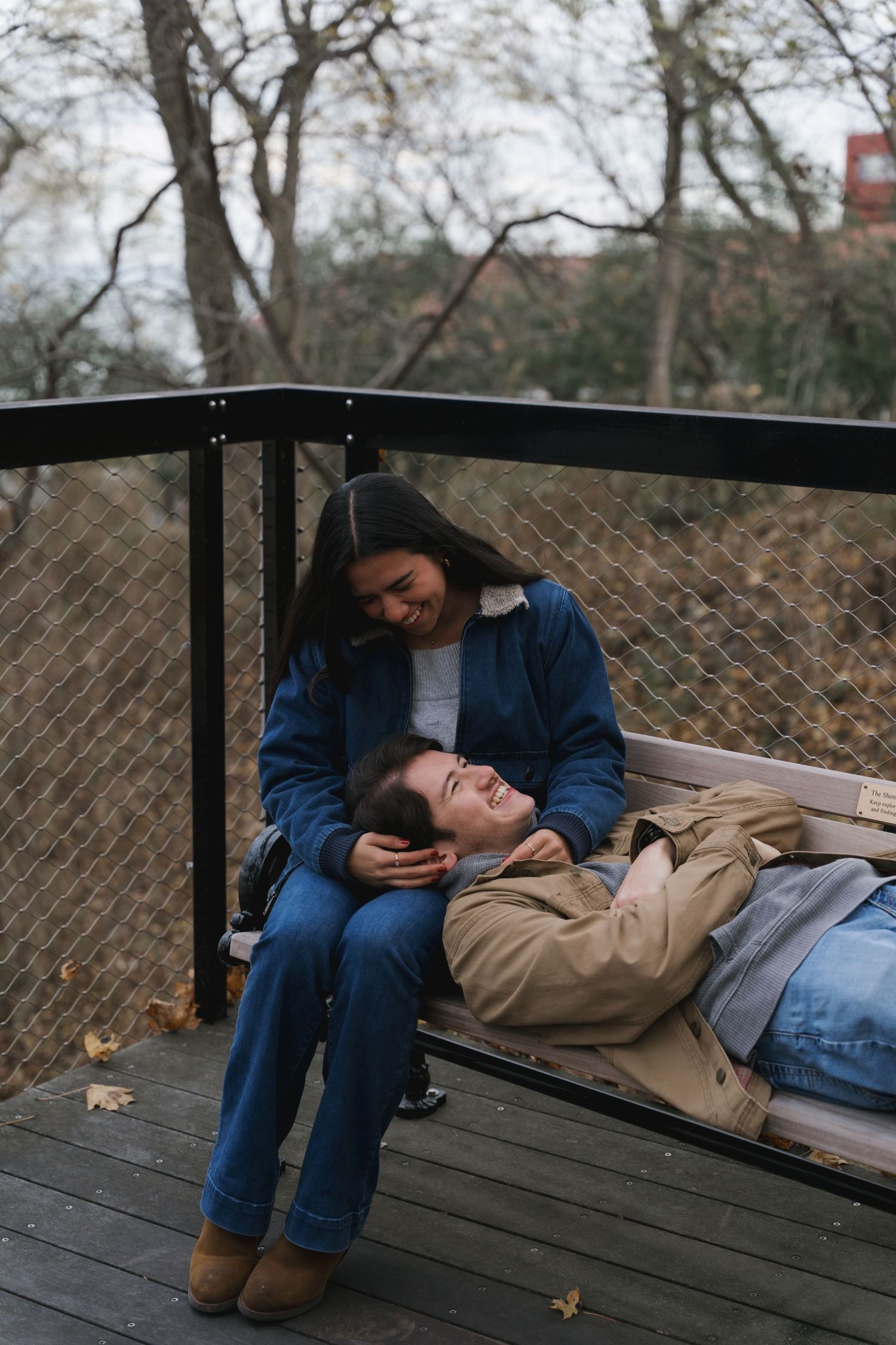 A woman smiles at her partner lying across her lap on an outdoor bench in autumn.
