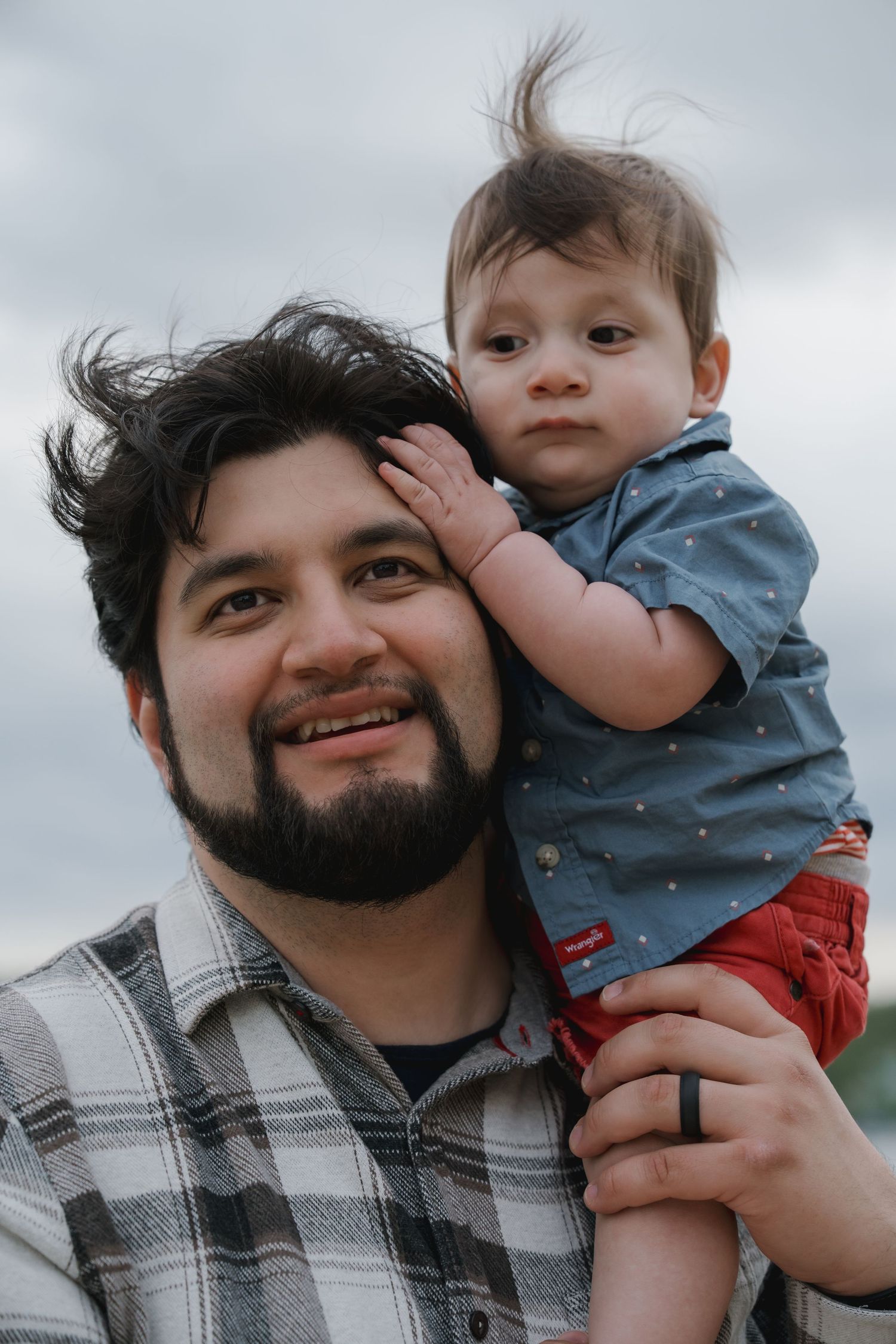 A bearded man smiles while holding a baby in a blue shirt outdoors under a cloudy sky.