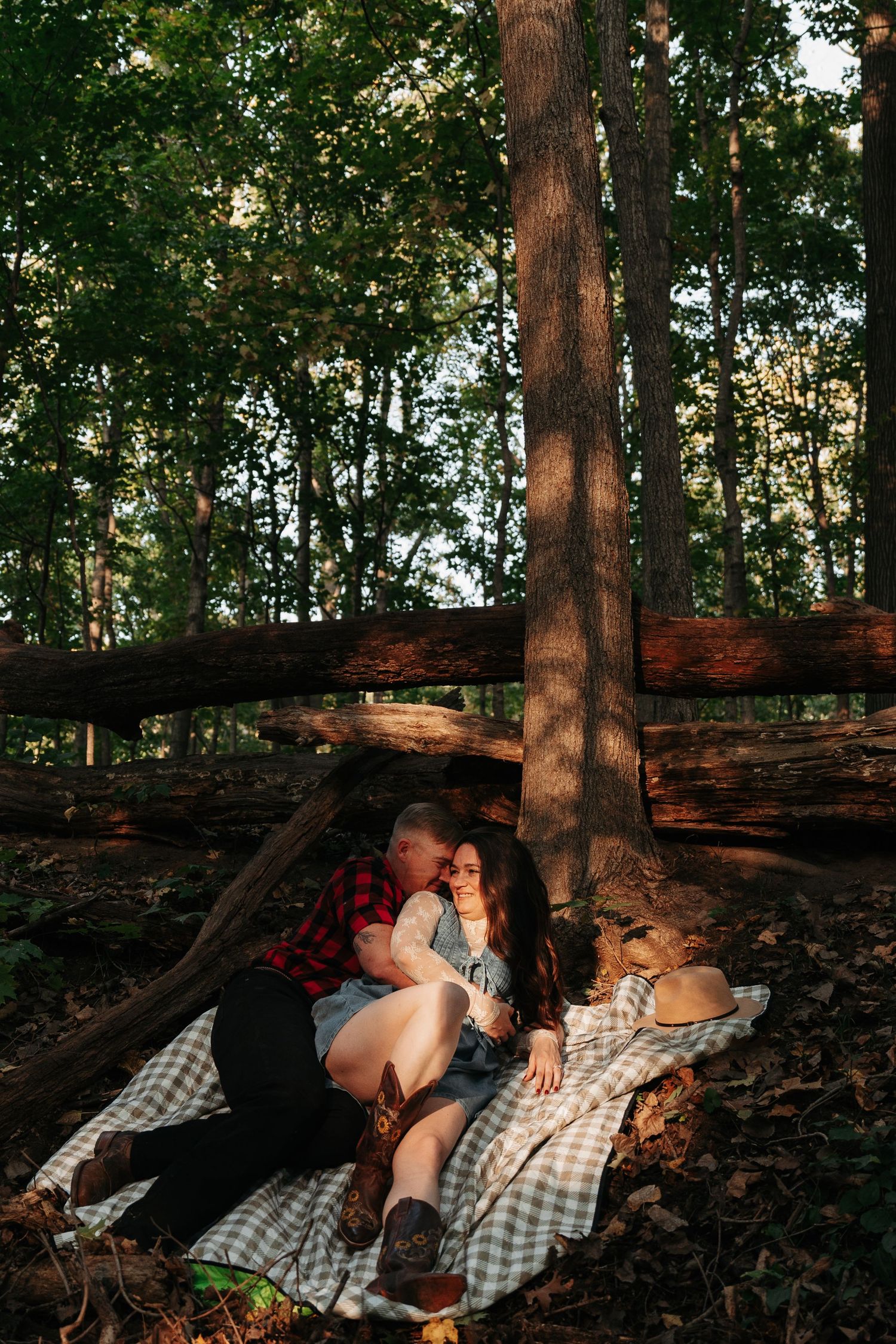 Couple cuddling on a blanket against a tree in a sun-dappled forest setting.