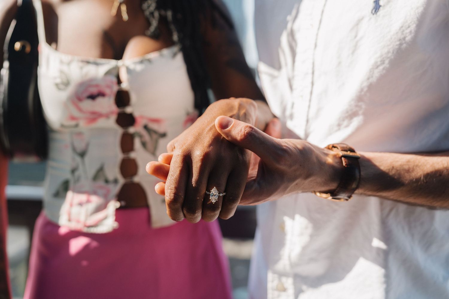 Close-up of couple holding hands showing diamond marquise cut engagement ring in sunlight.