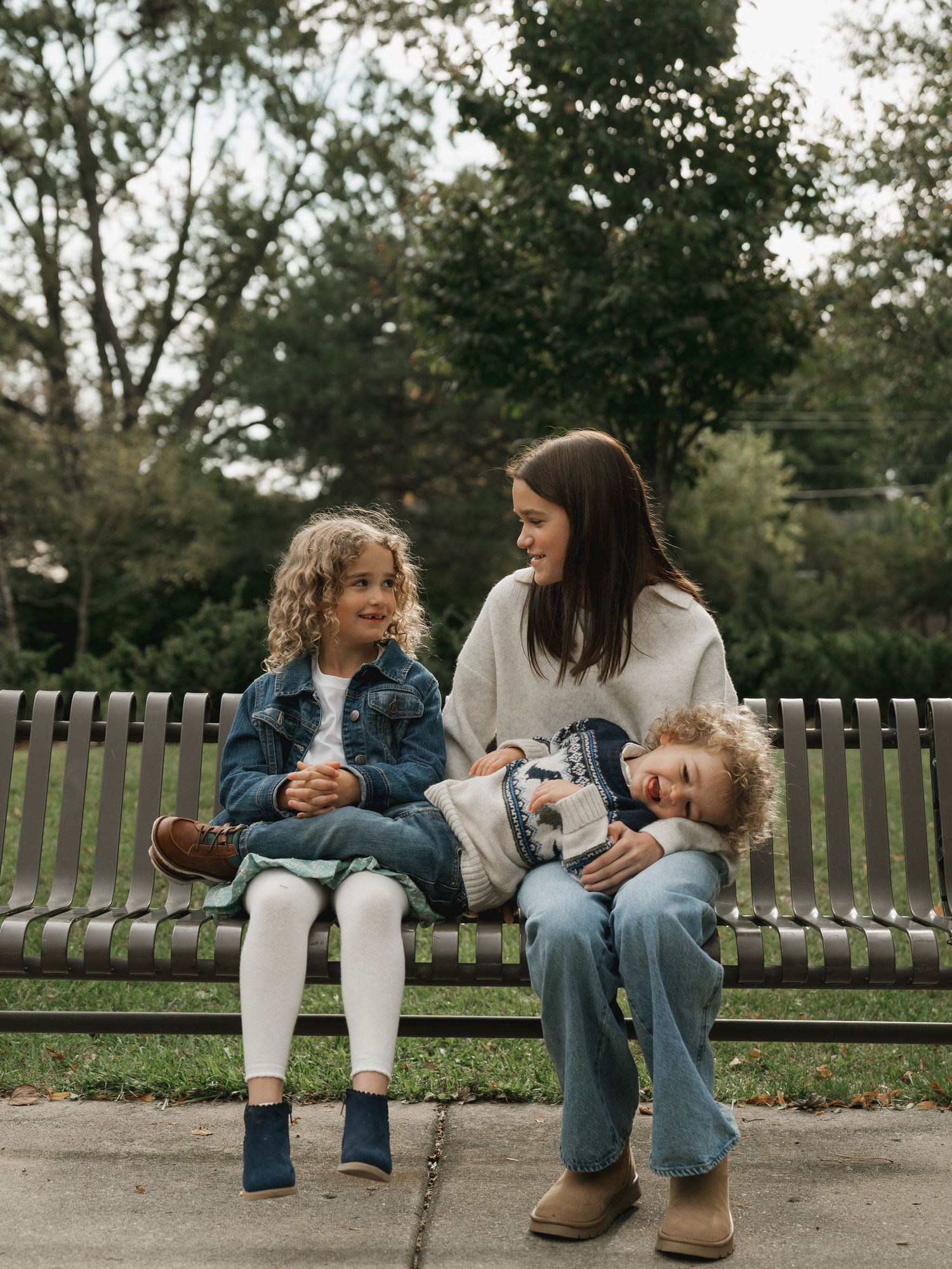 Siblings sitting on park bench smiling at curly-haired sister while younger child rests on her lap.