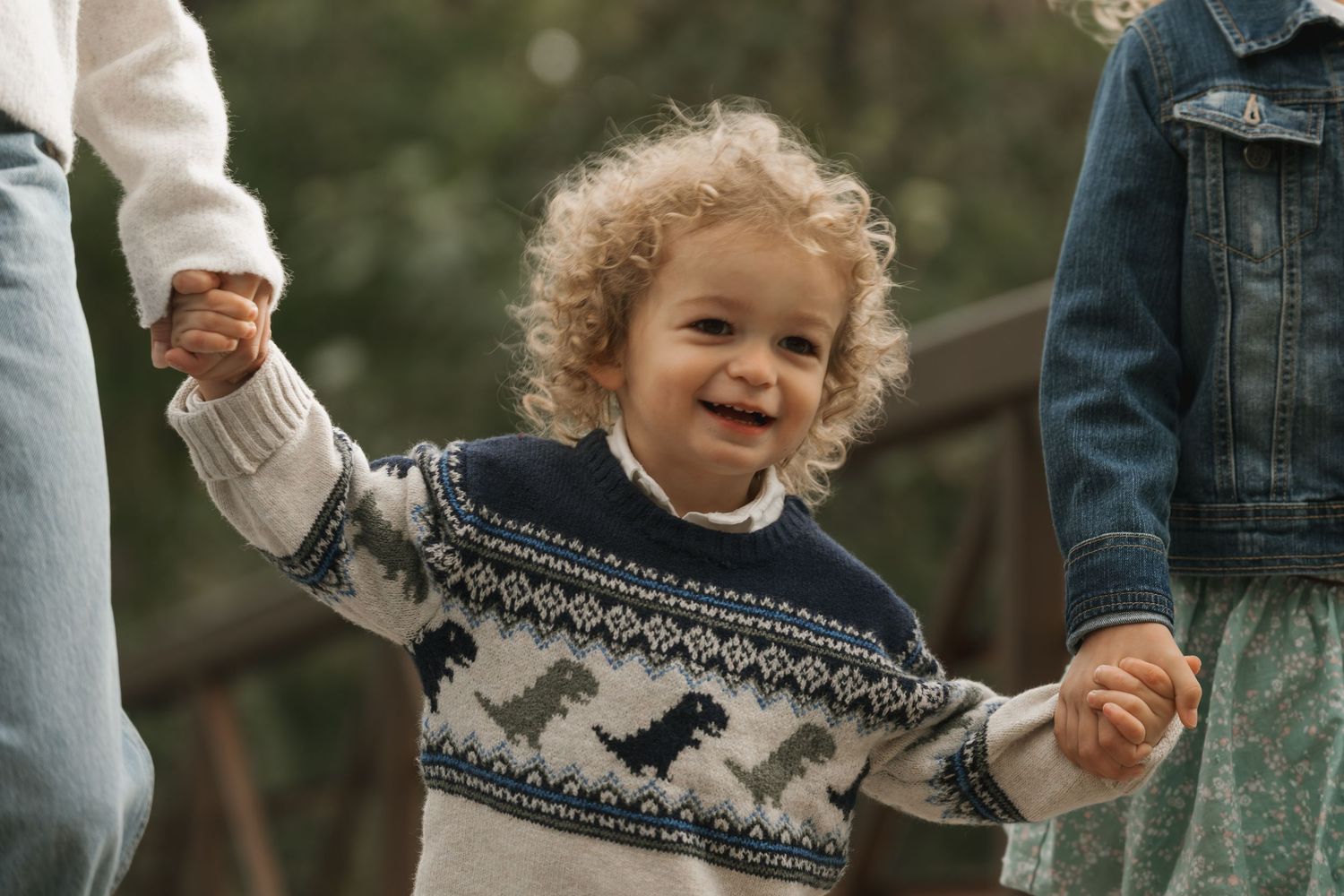 Happy toddler in dinosaur sweater holding hands with family outdoors.