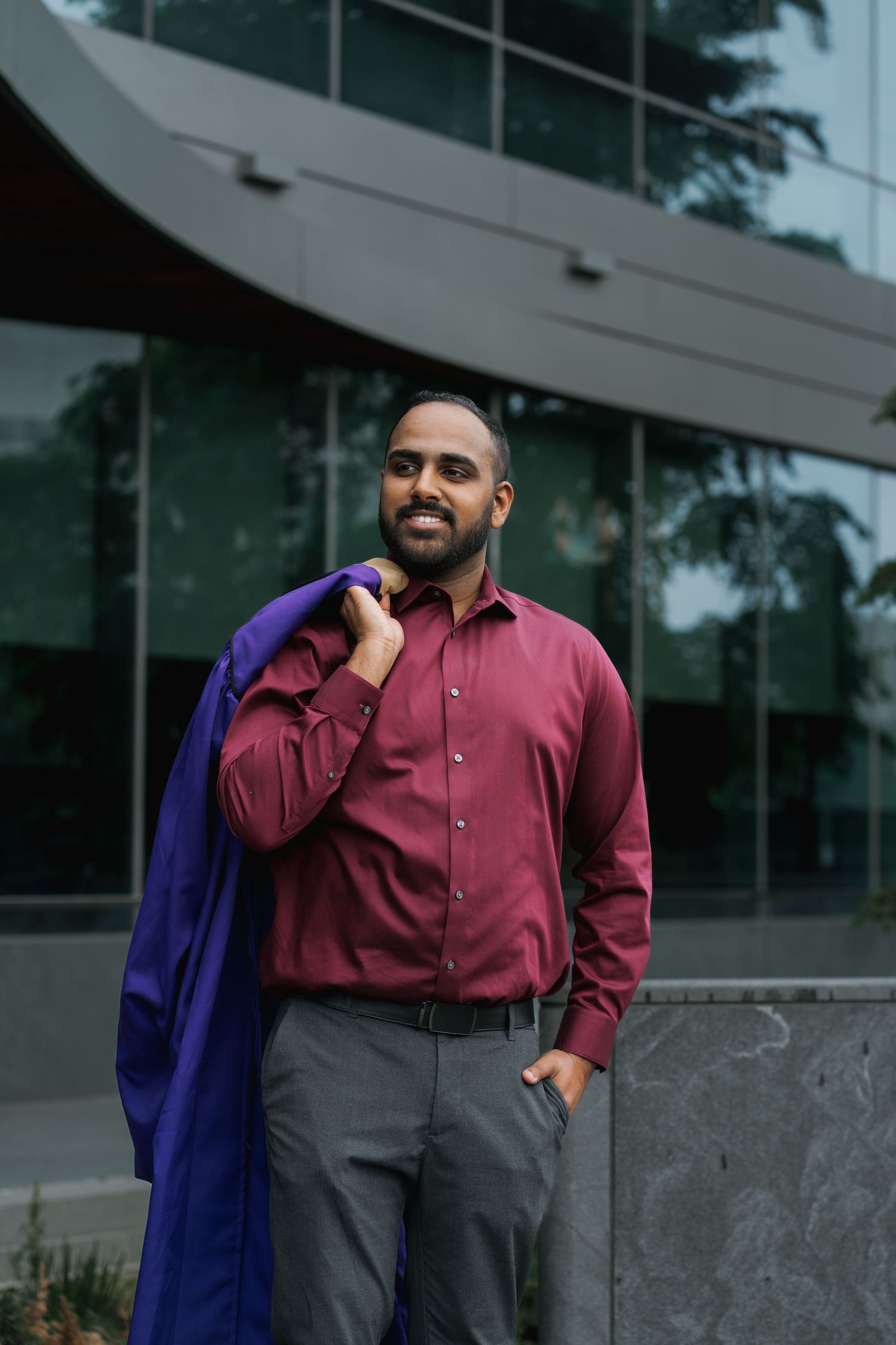 A confident man in a maroon shirt carries a purple graduation gown over his shoulder outdoors.