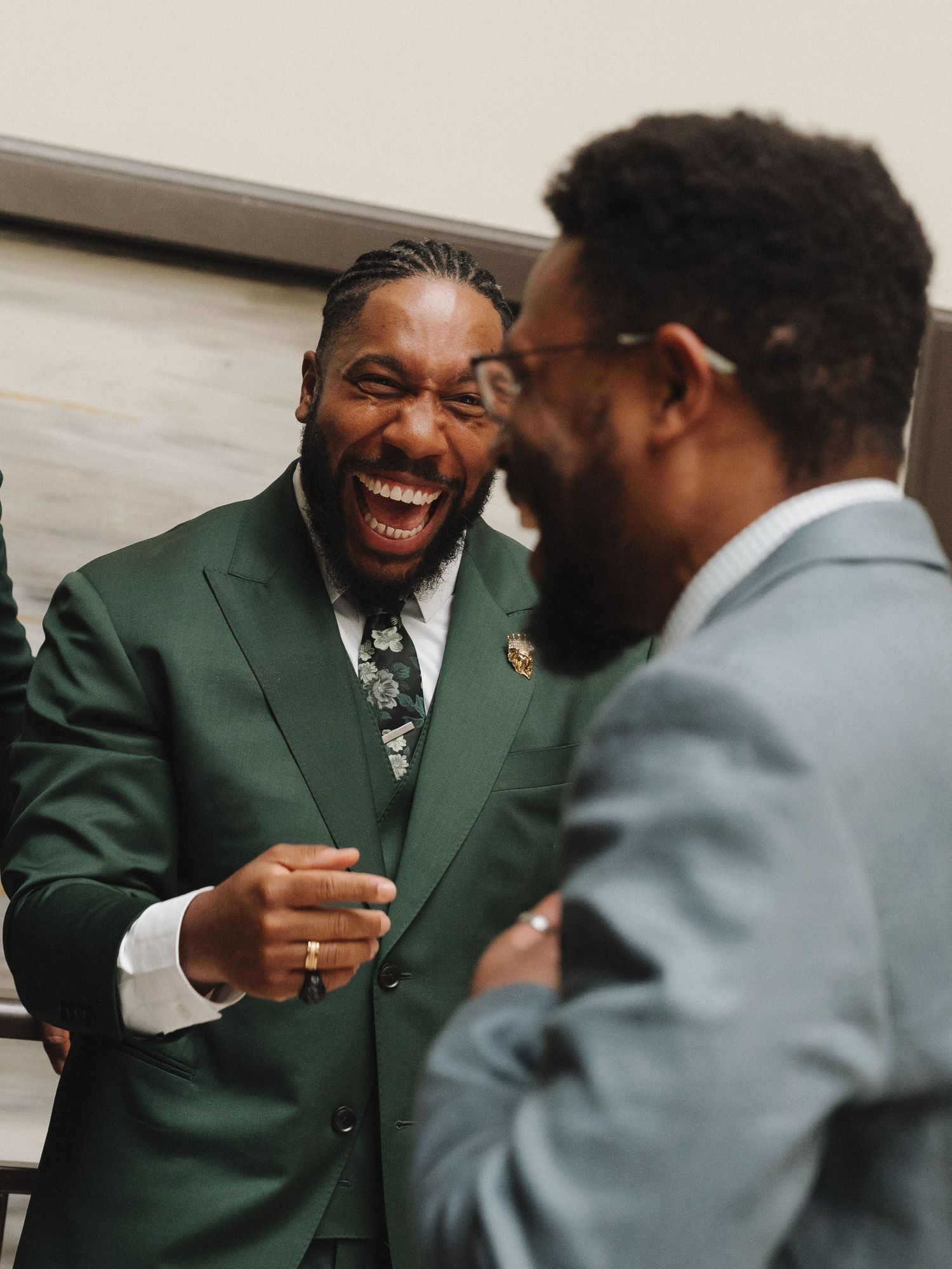 Two men in suits laughing joyfully at a formal event, one wearing a green suit with floral tie.