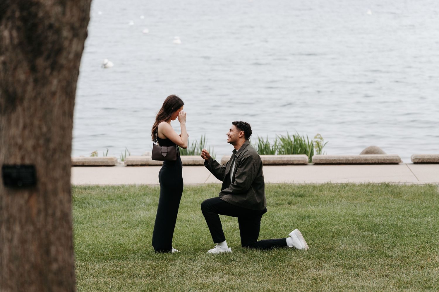 Man proposes to woman on one knee by lakeside on green grass in romantic outdoor engagement moment.