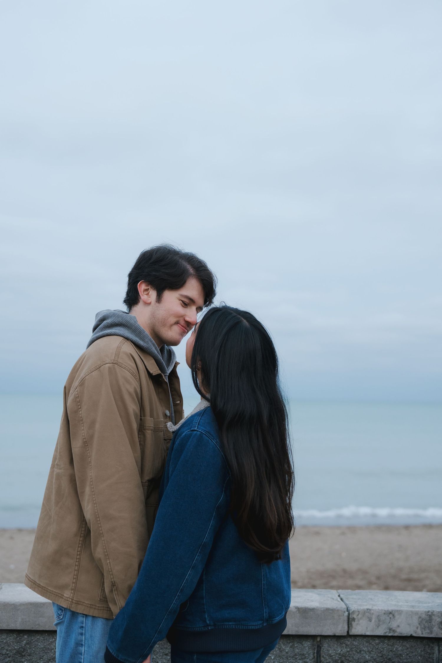A couple leans in for a kiss while standing near a stone wall at the beach.