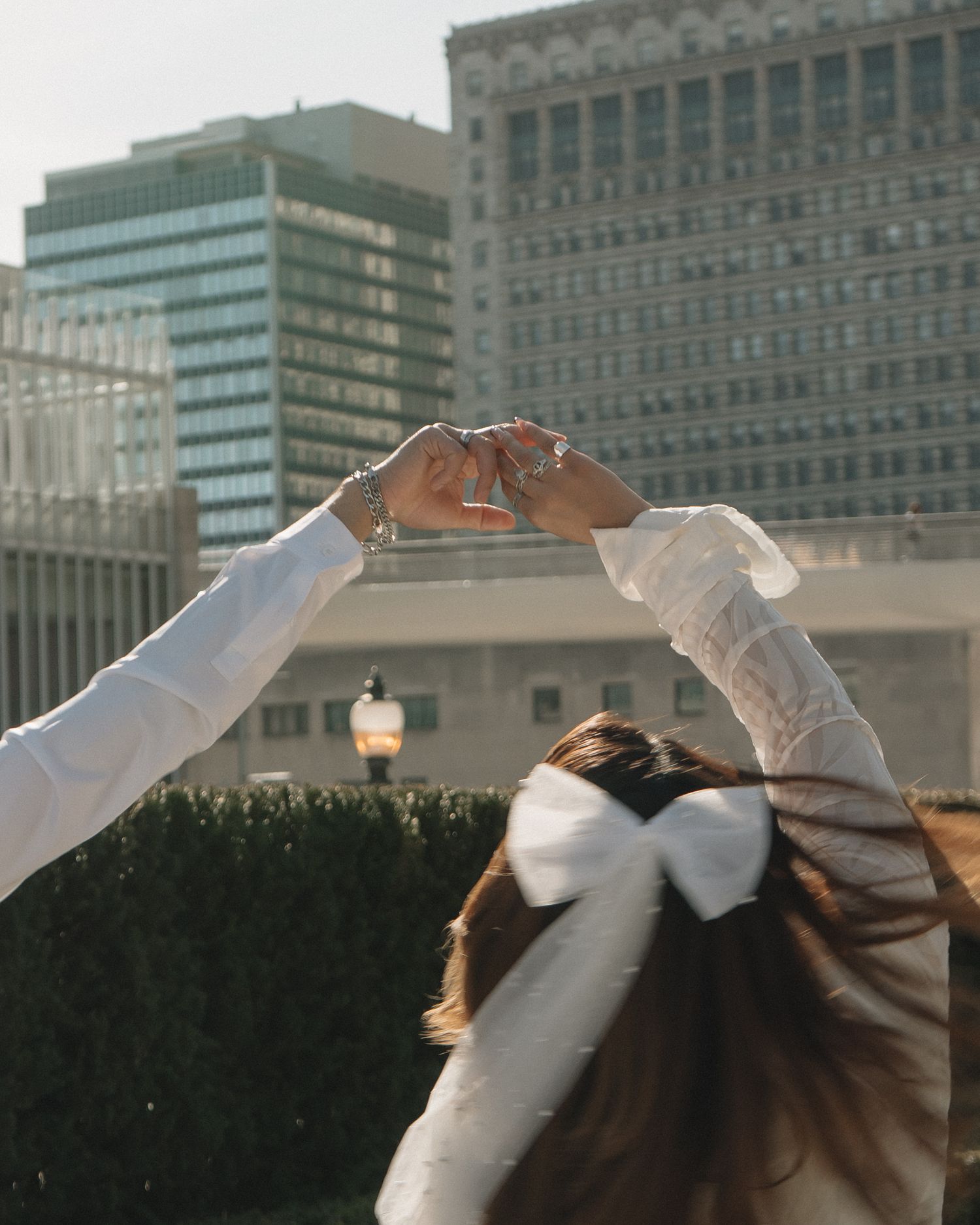 Woman in white dress with large bow raises hands to form a heart shape against city skyline.