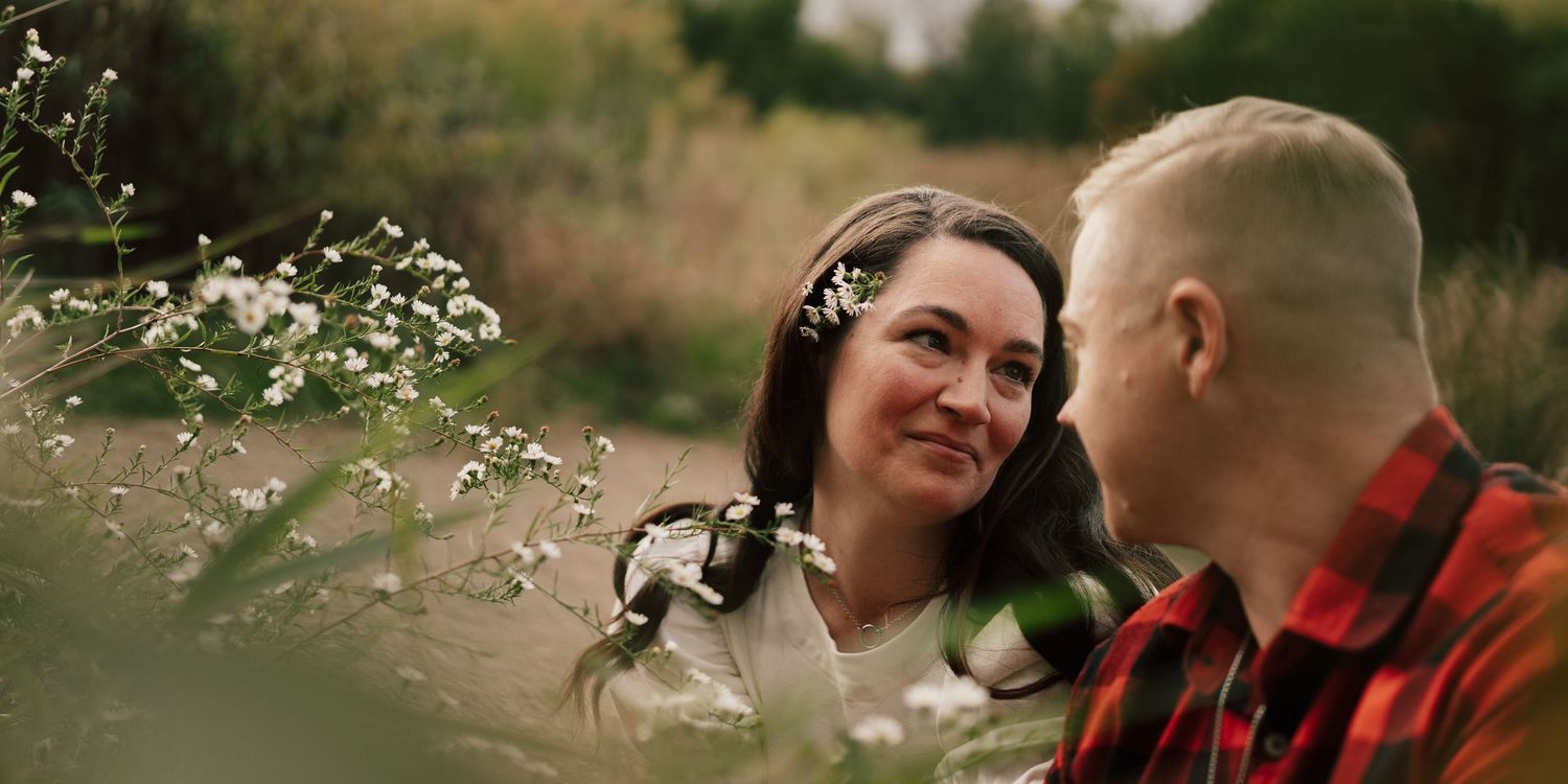 Smiling woman with a flower in her hair gazes at her partner among wildflowers in a field.