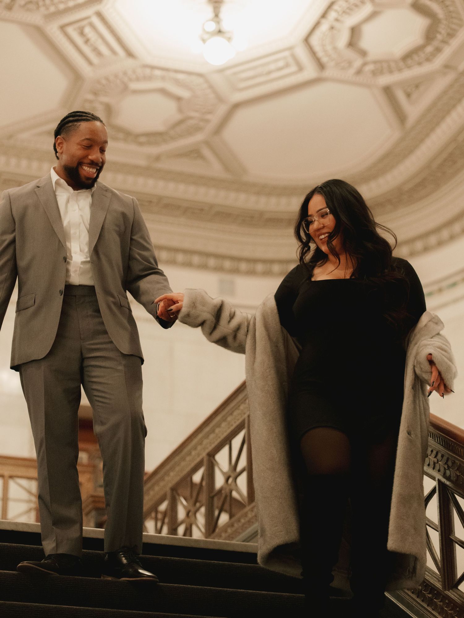 Happy couple walking down grand staircase, man in gray suit holding womans hand.