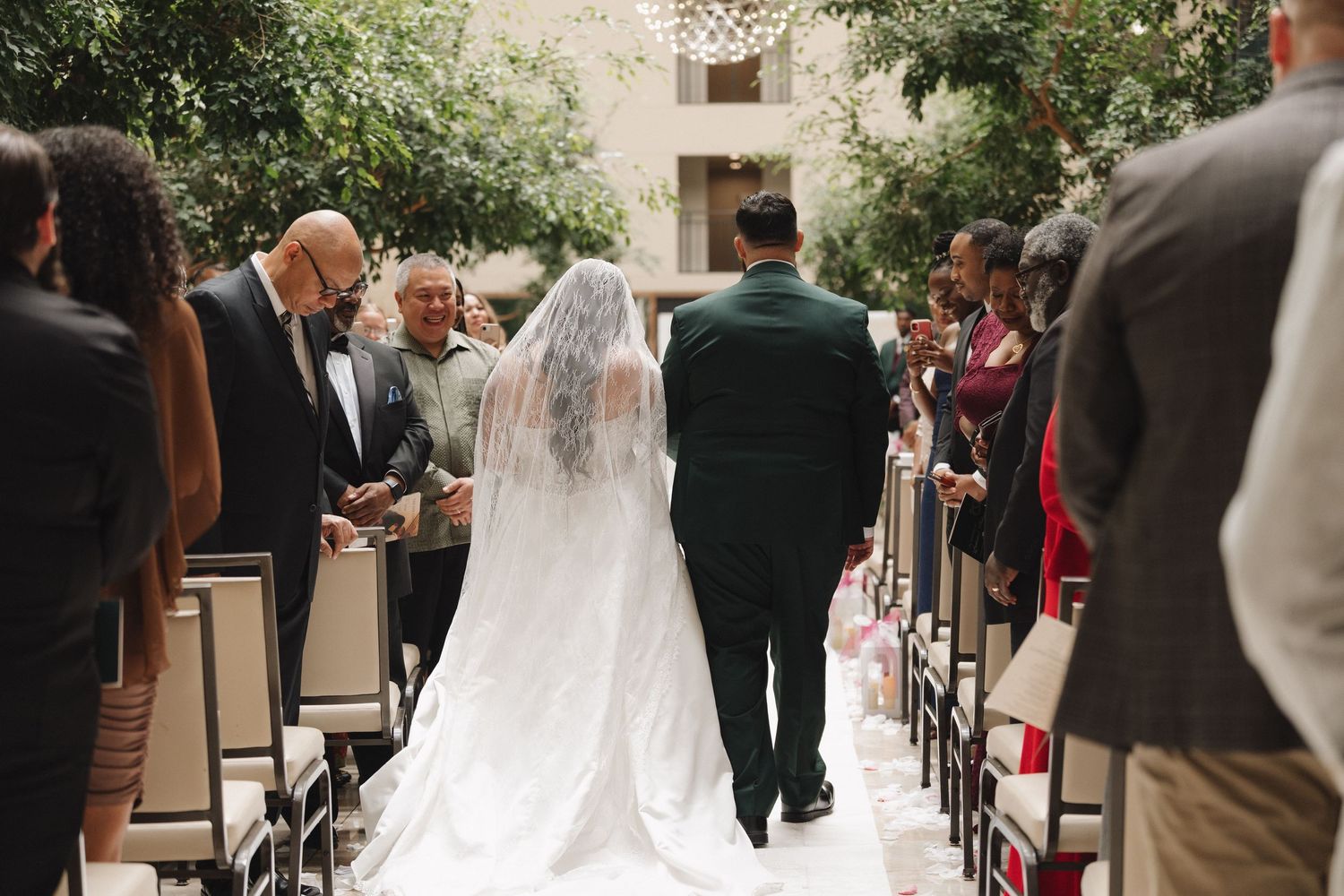 Bride in white gown and veil walks down the aisle escorted by two men at an indoor wedding ceremony.