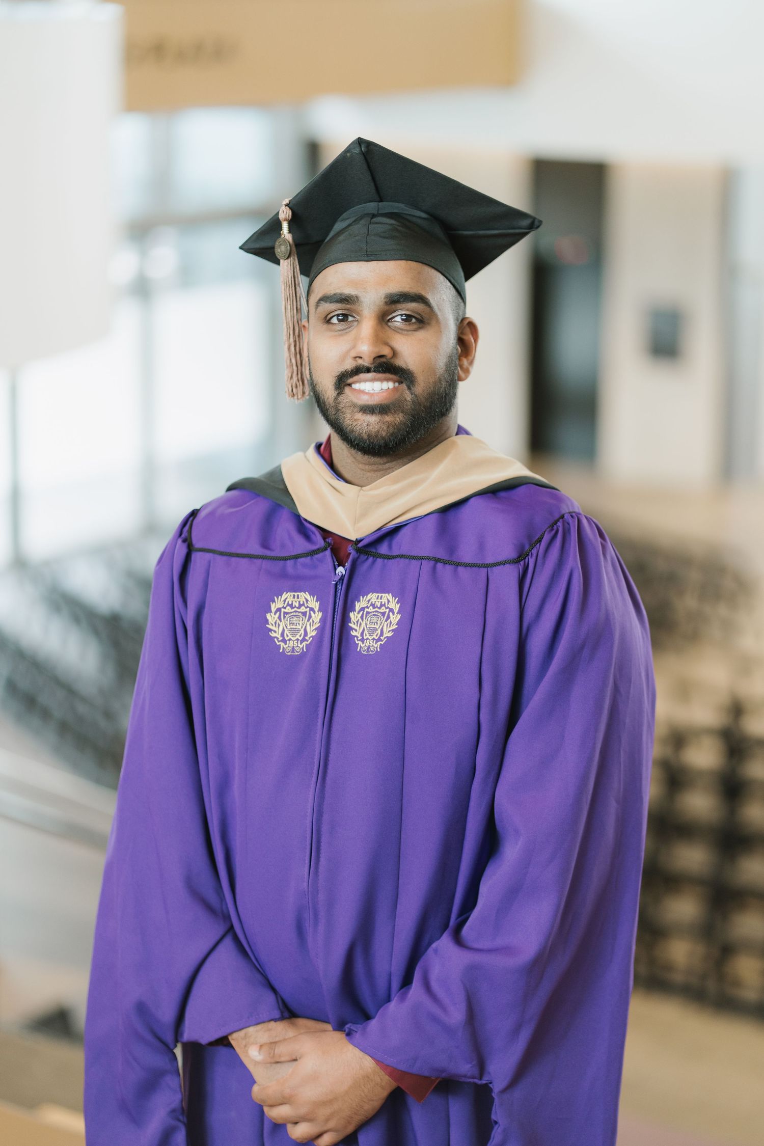 A smiling man in a purple graduation gown and black mortarboard cap poses indoors at graduation.