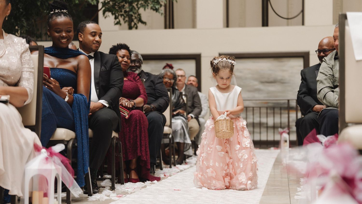 A young flower girl in a pink dress walks down the wedding aisle during an elegant indoor ceremony.
