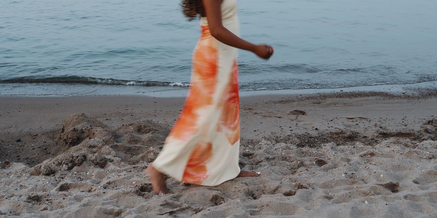 Woman in flowing orange and white tie-dye maxi skirt walking barefoot on sandy beach near ocean waves.