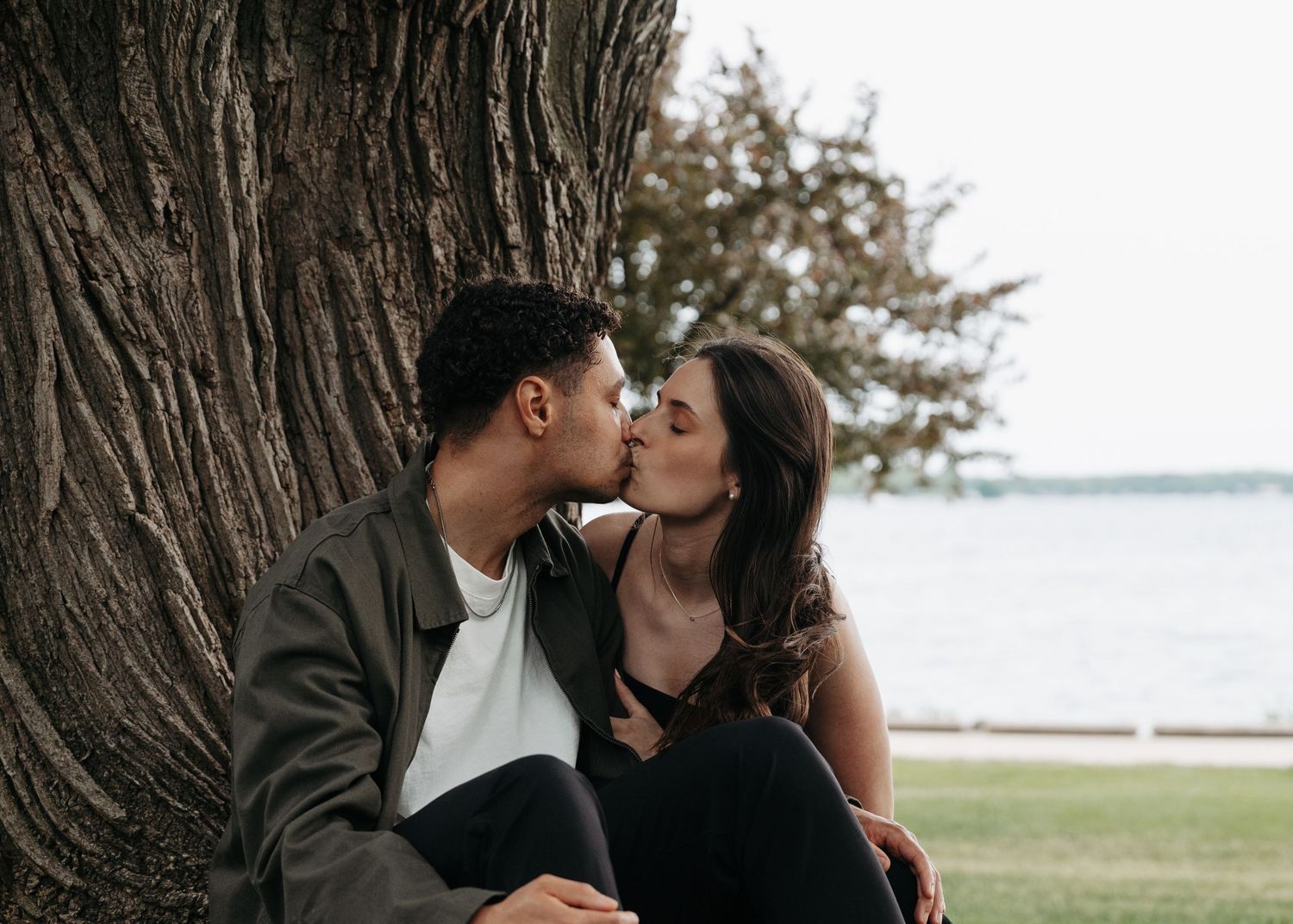Couple sharing a romantic kiss while sitting against a large tree near a waterfront on a cloudy day.