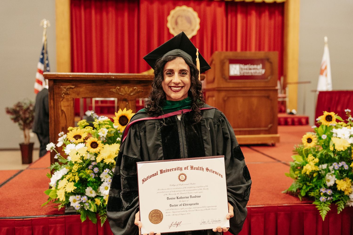 Woman in graduation cap and gown holding diploma at National University of Health Sciences commencement ceremony.