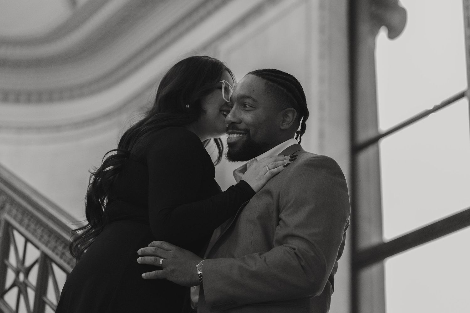 Black and white romantic photo of couple embracing and almost kissing near architectural columns.