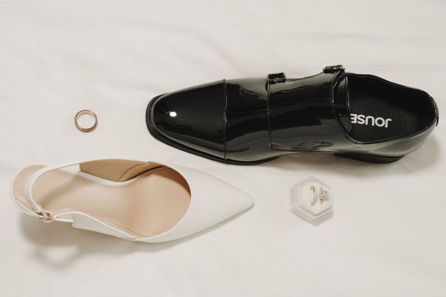 Elegant wedding flat lay with white bridal heels, black patent leather dress shoes, gold wedding bands, and a diamond ring on white fabric.