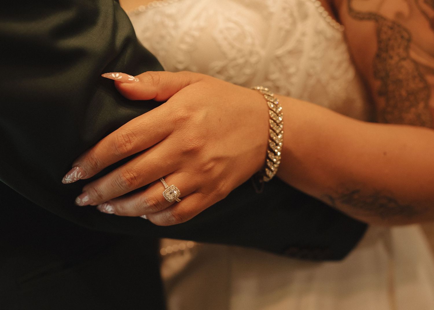 Bride's hand with diamond ring and gold bracelet resting on groom's suit, showing elegant long nails and tattoo.