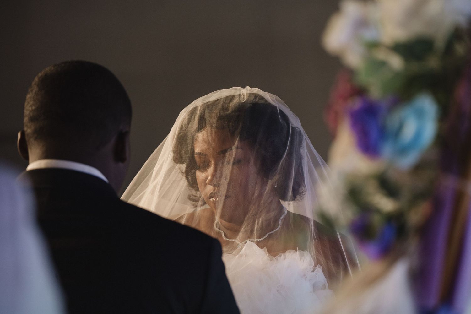 Bride in white veil bows head during intimate wedding ceremony moment.