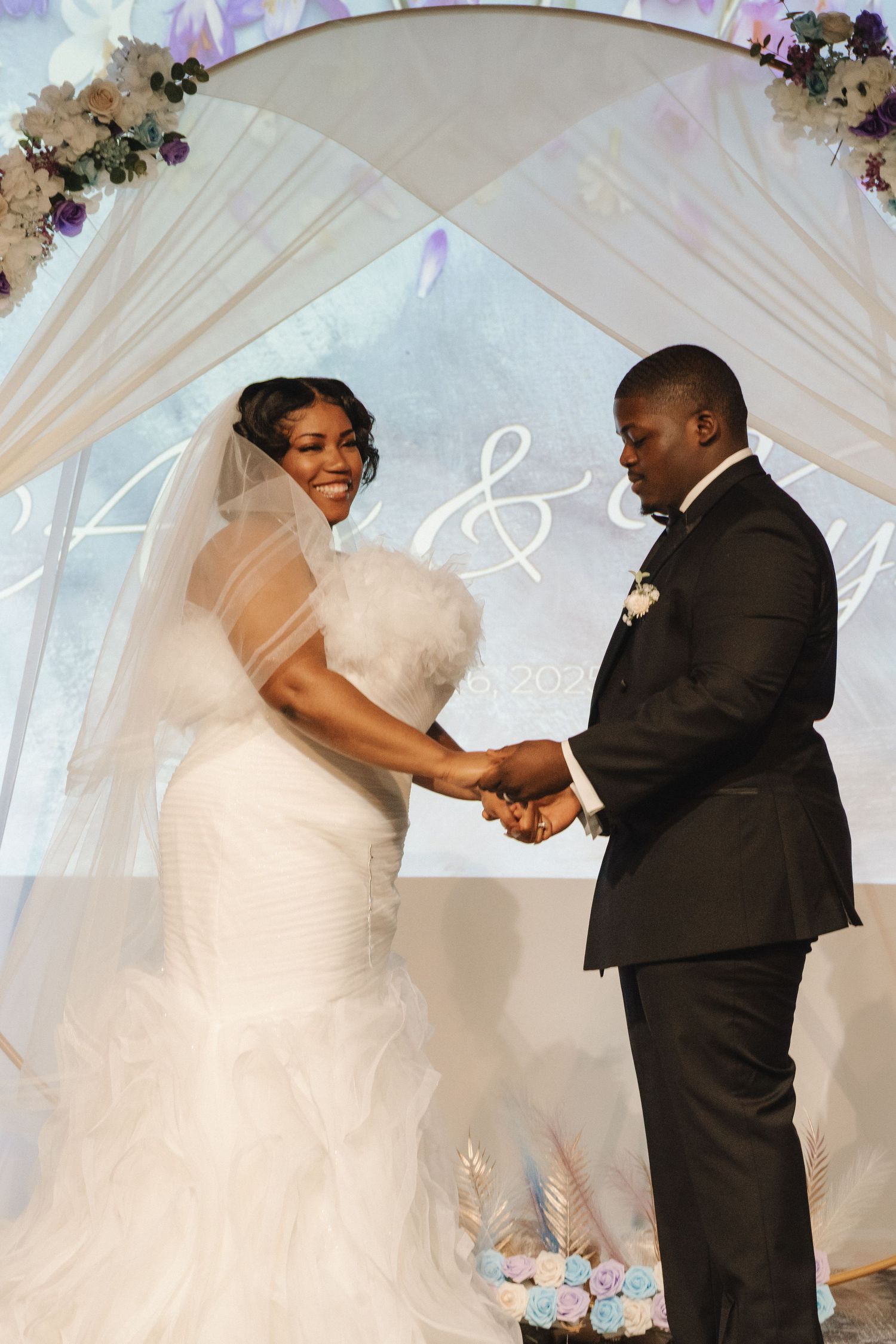 Bride and groom smile holding hands under floral arch at wedding altar.
