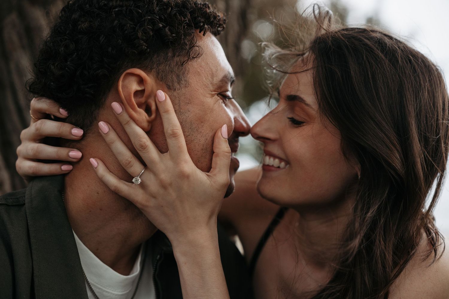 Couple sharing a romantic moment, woman smiling with engagement ring visible, noses touching tenderly outdoors.