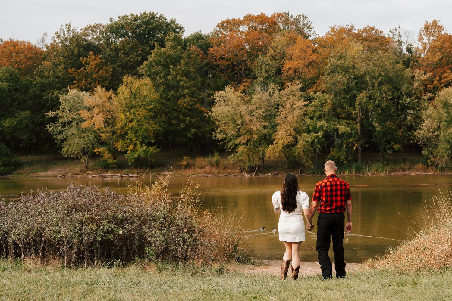 Couple holding hands facing a serene autumn pond surrounded by colorful fall foliage trees in a park.