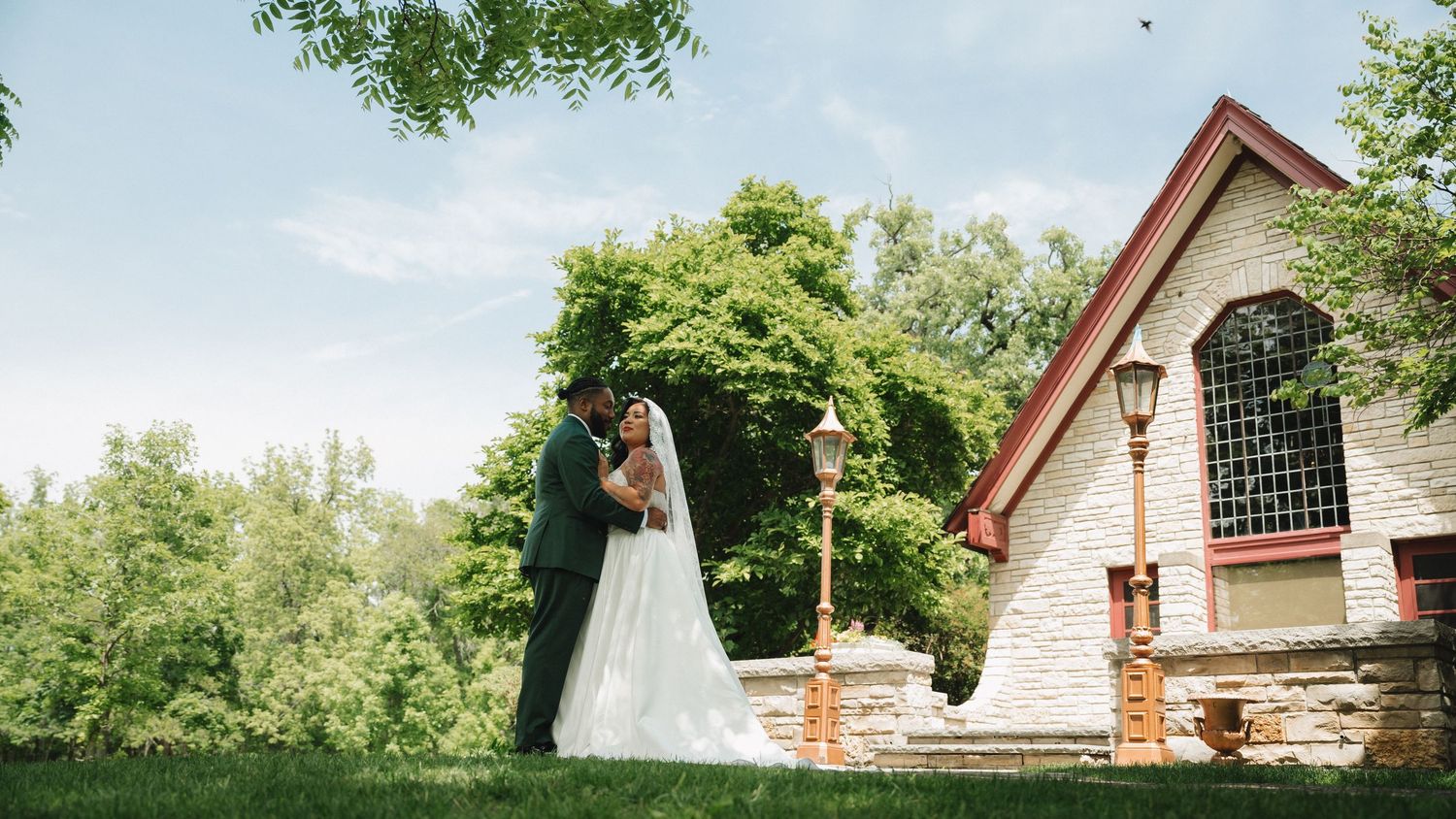 A bride and groom share a romantic kiss on lush green grounds beside a charming stone chapel on their wedding day.