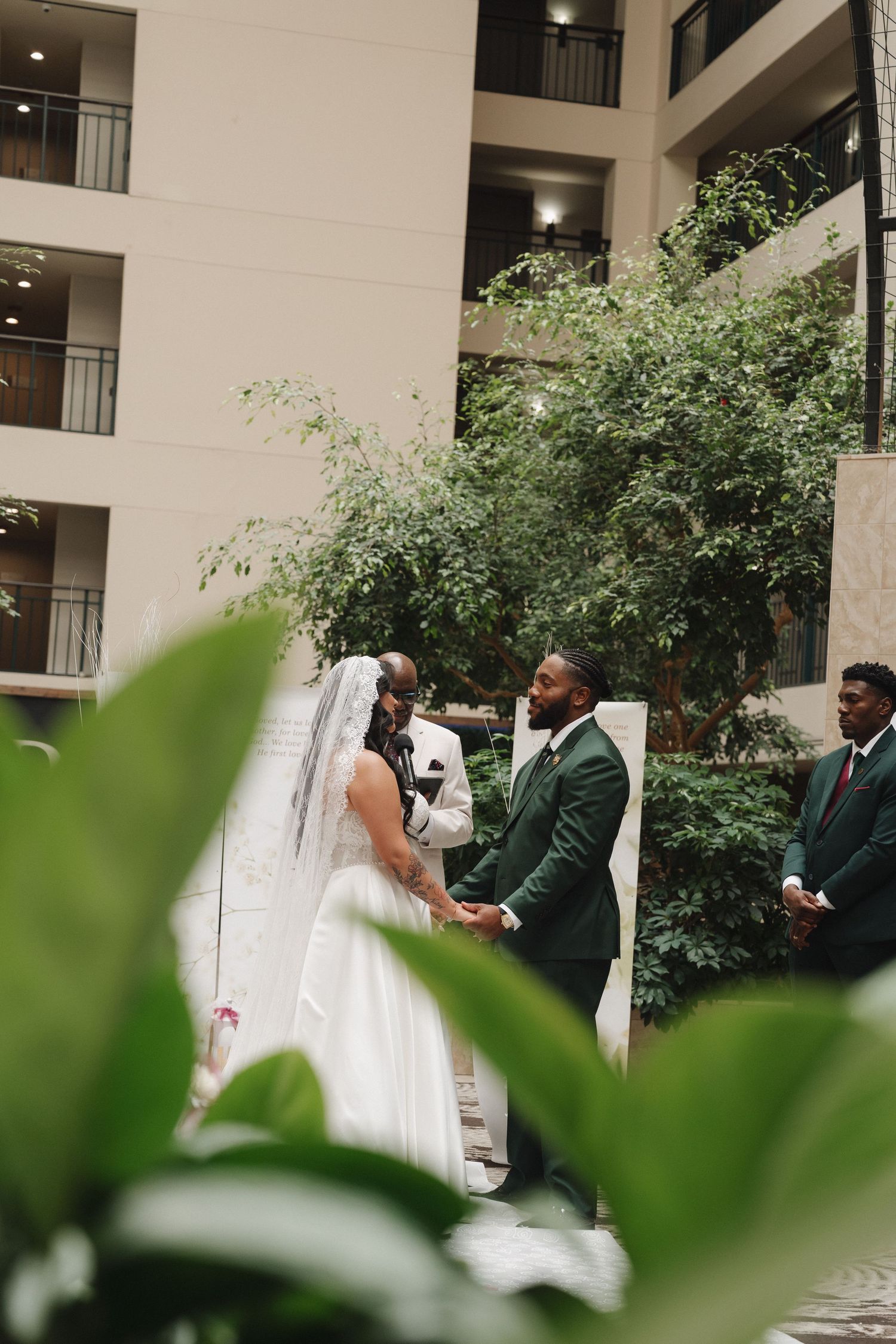 Groom in dark green suit holds hands with bride in white gown during indoor wedding ceremony surrounded by lush greenery.