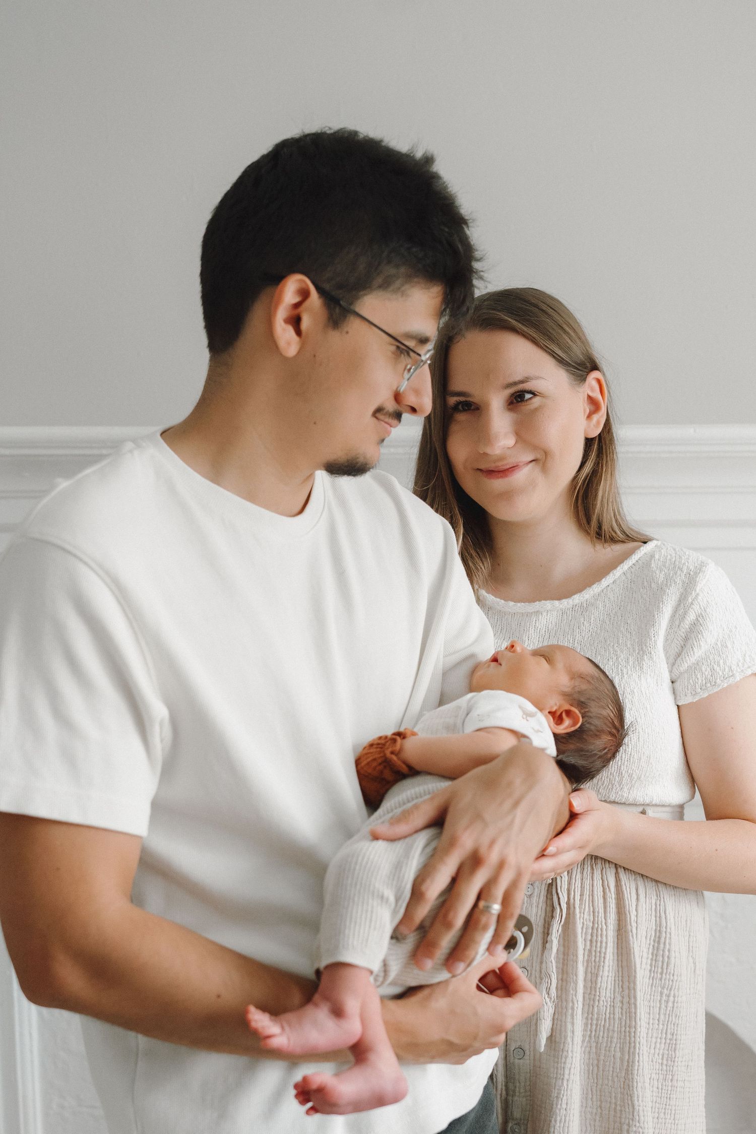 Young couple in white clothing tenderly holding their newborn baby against a soft gray background.