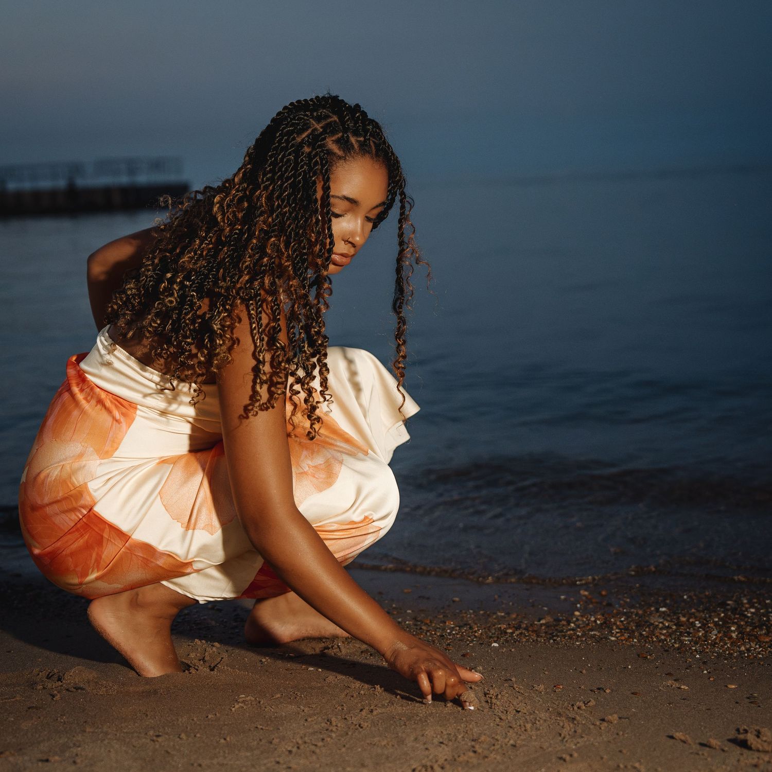 A young woman with braided hair in an orange dress crouches on a sandy beach at dusk, touching the sand.