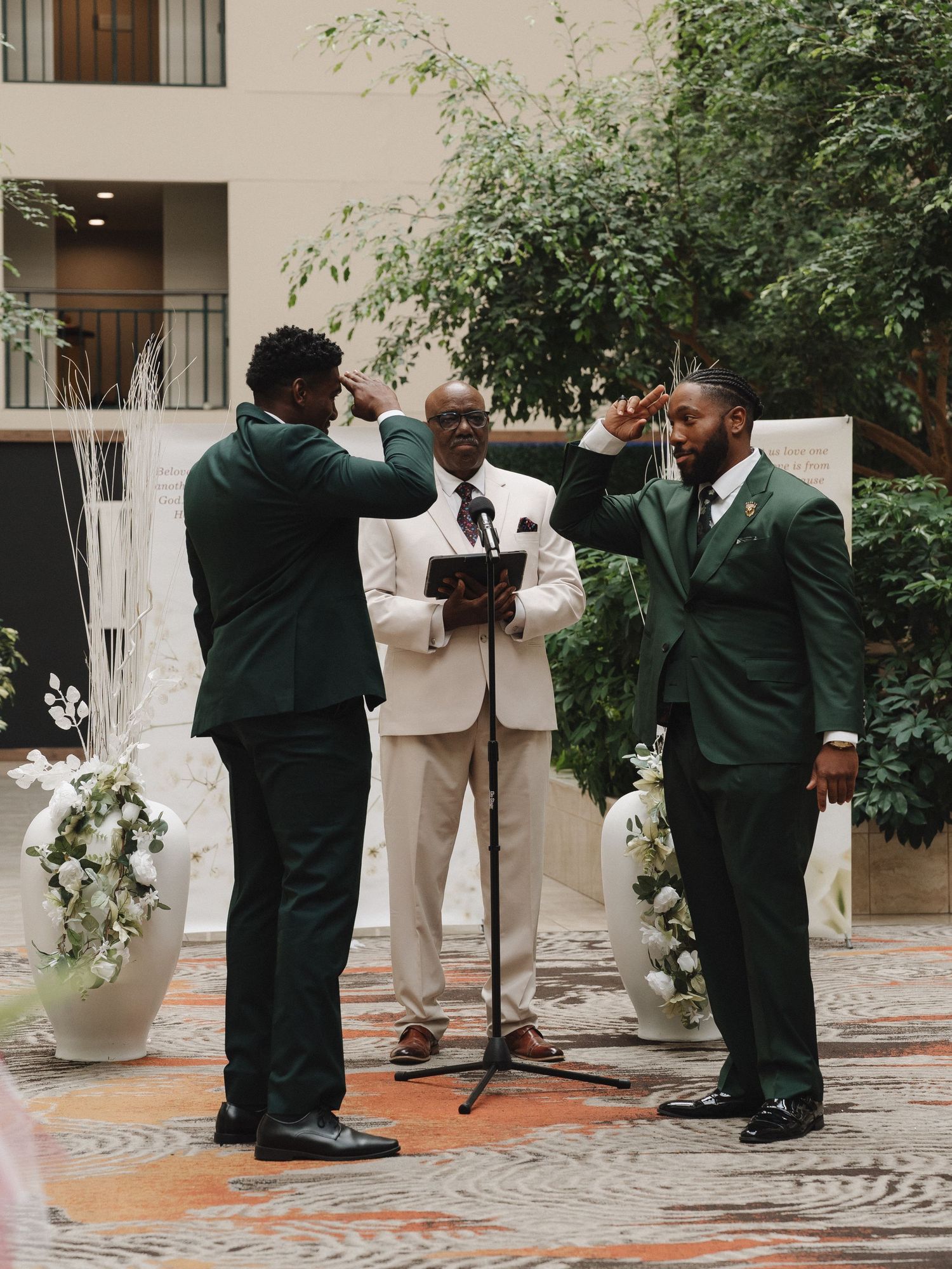 Two grooms in dark green suits salute each other at indoor wedding ceremony with officiant and floral decor.