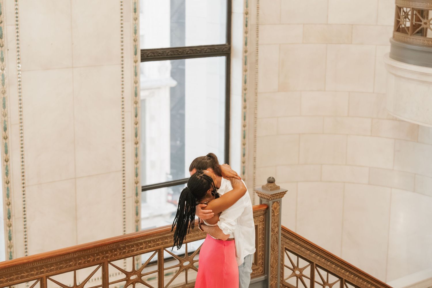 Couple hugging near a tall window on a decorative staircase in a historic building.