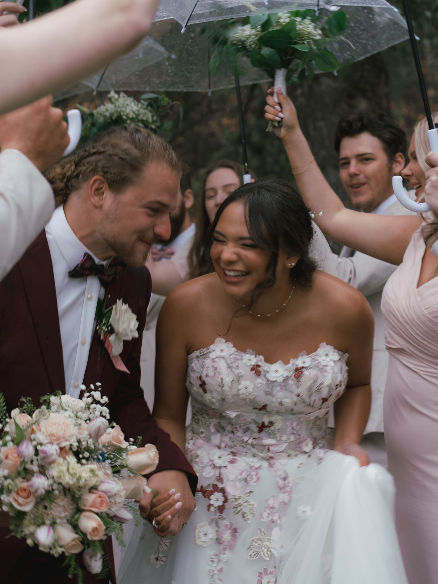 Smiling bride and groom walk through cheering wedding party guests holding umbrellas in the rain.