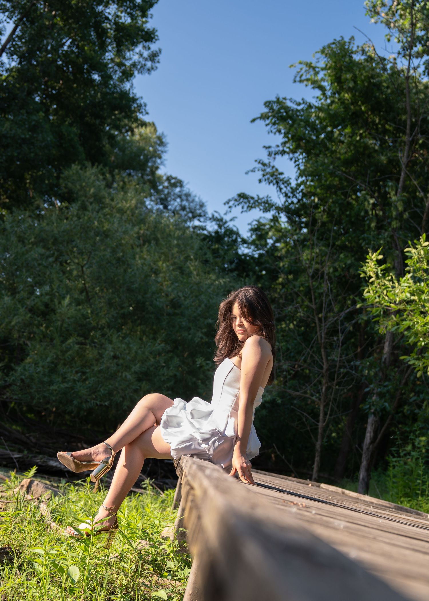 Young woman in white sundress sitting on wooden fence rail in sunny green outdoor setting.