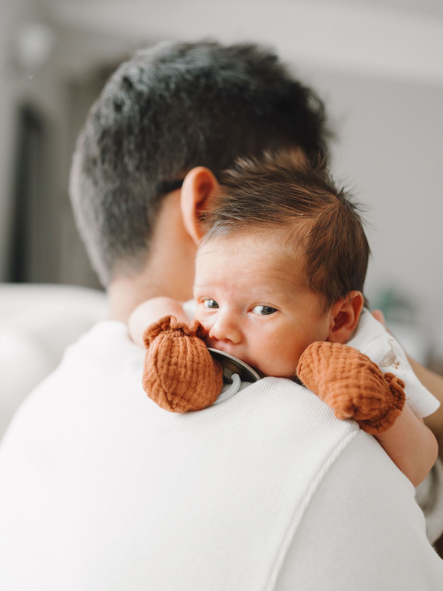 Newborn baby with brown knit mittens resting on father's shoulder looking over his back.