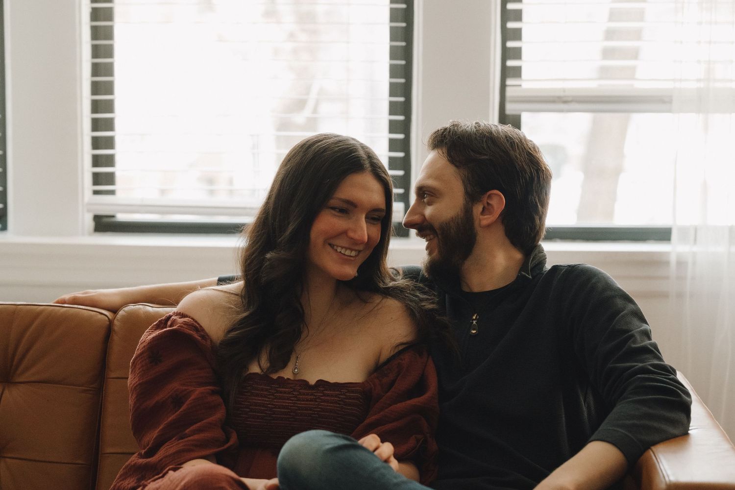 A smiling couple sitting together on a brown leather couch, sharing a warm and loving moment near a window.