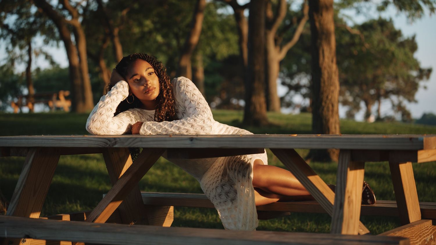 Young woman in white lace dress poses at outdoor picnic table surrounded by trees in warm golden sunlight.