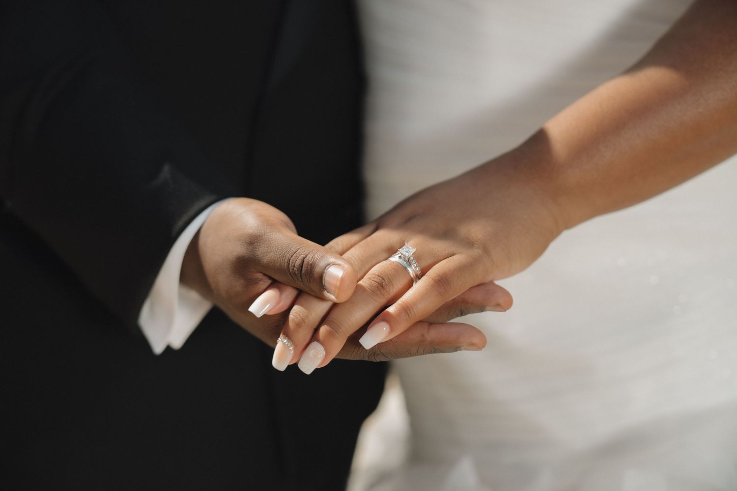 Soft, natural light illuminates a bride and groom's hands in a moment of quiet connection, highlighting the elegant details of their wedding