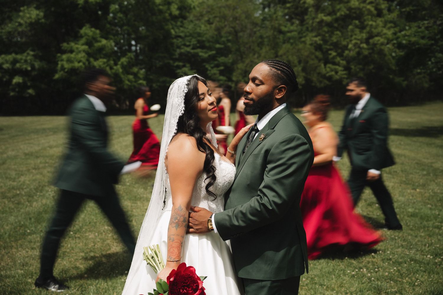 Bride and groom share a romantic moment outdoors, with wedding party in red and green attire blurred in motion behind them.