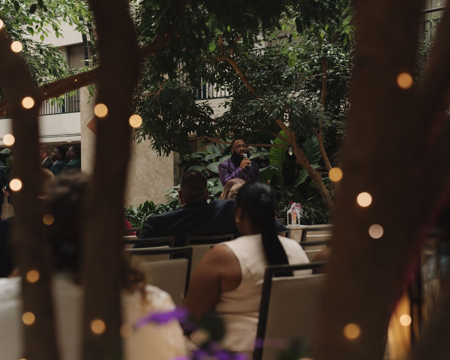 Intimate wedding ceremony moment captured through trees with warm string lights in a lush hotel atrium.