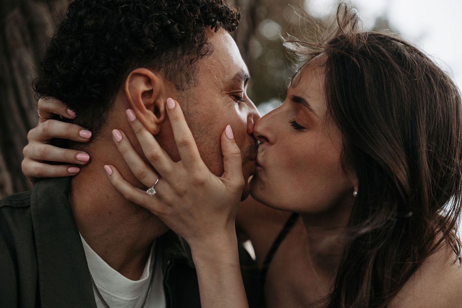 Romantic couple sharing a passionate kiss outdoors, woman wearing a ring, embracing tenderly near a tree.