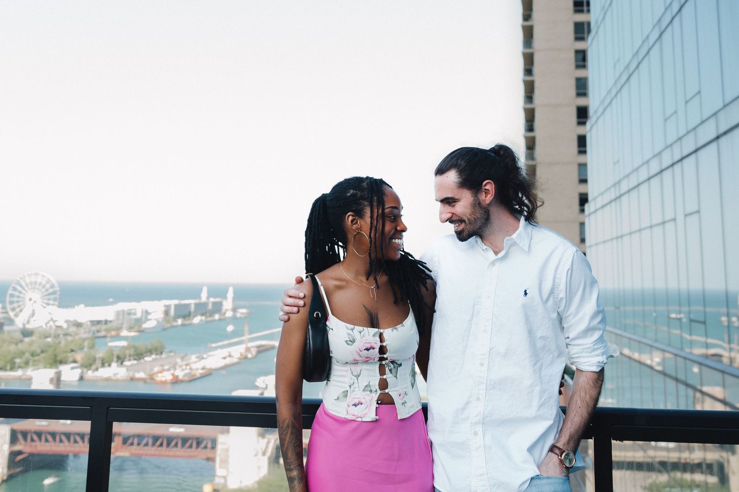 Happy interracial couple smiling at each other on rooftop balcony with city skyline behind.