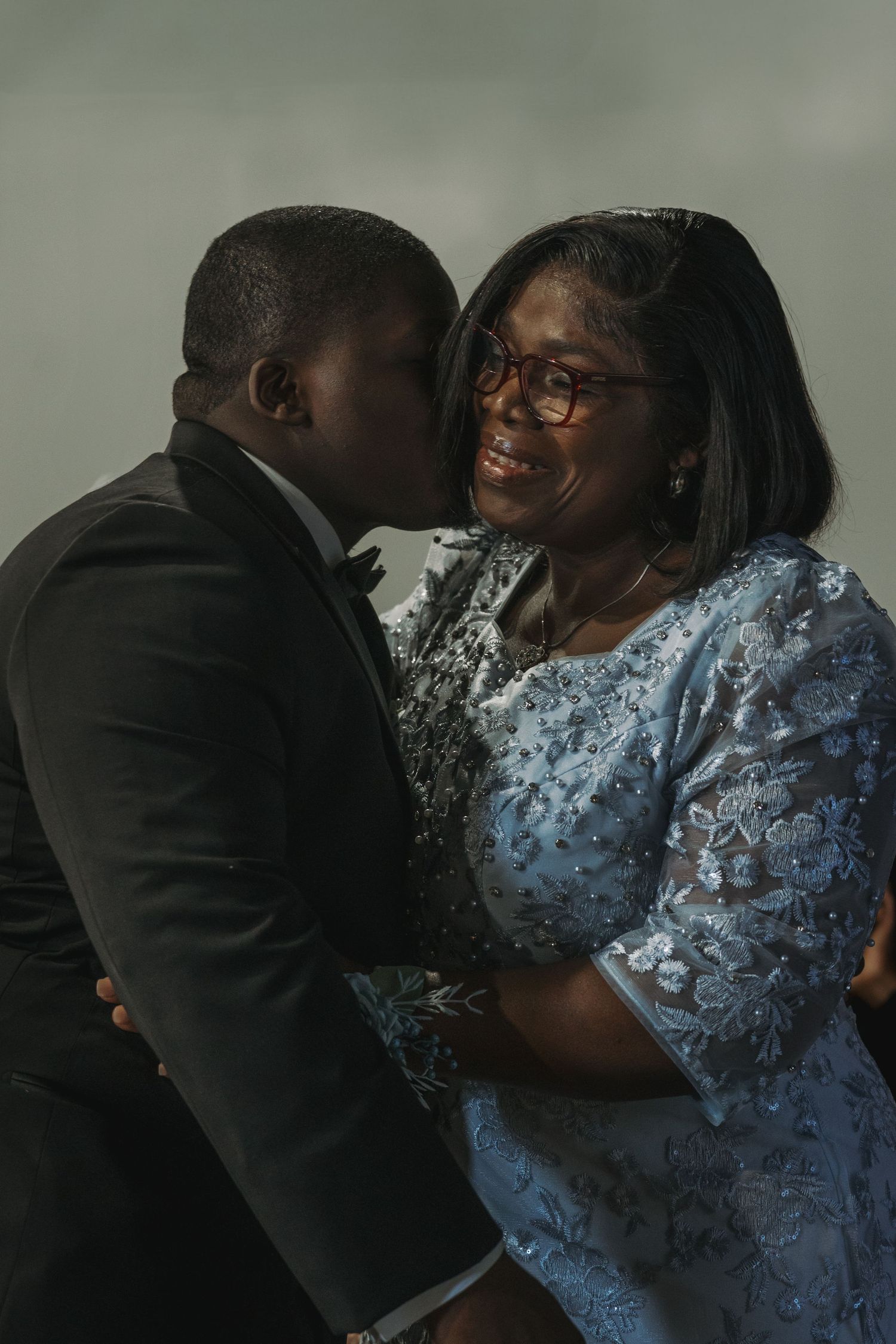 Groom in black suit kisses the cheek of his mother wearing a blue lace dress.