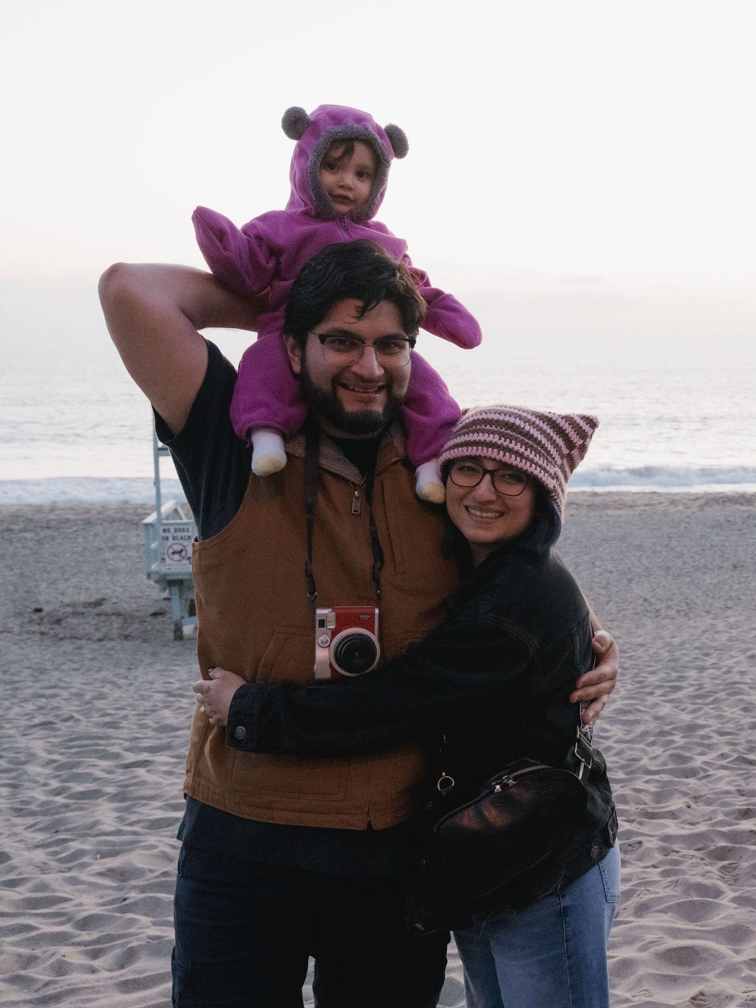 Family of three poses on beach at dusk, man carries toddler in purple bear costume on shoulders while woman hugs him.