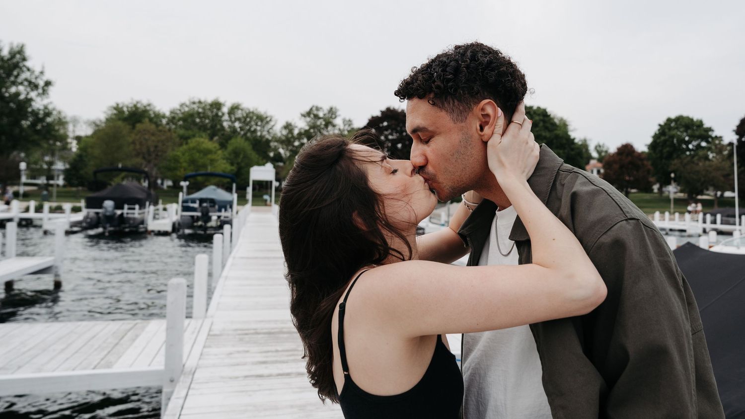 A couple shares a romantic kiss on a waterfront dock with boats and lush green trees in the background.